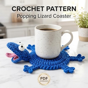 May include: A blue crocheted lizard-shaped coaster with a pink tongue and white eyes, featuring a speckled white ceramic mug placed on top. The text CROCHET PATTERN Popping Lizard Coaster and a gold PDF DOWNLOAD badge are visible in the image.