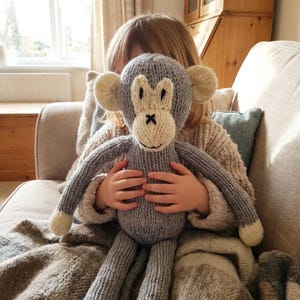 Può includere: Un peluche a forma di scimmia lavorato a maglia a mano con corpo grigio e dettagli color crema su orecchie, mani e viso. La scimmia presenta occhi neri ricamati, un naso cucito e una semplice linea per la bocca, realizzato in filato morbido.