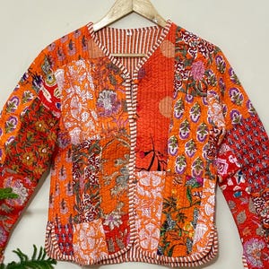 Puede incluir: Chaqueta patchwork con estampados florales en tonos naranja y rojo. La chaqueta tiene mangas largas y cuello redondo. El diseño incorpora varios parches de tela, creando una prenda única y colorida. La chaqueta está colgada en una percha de madera.