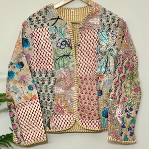 Puede incluir: Chaqueta patchwork con diversos estampados florales y geométricos en tonos beige, rojo y azul. La chaqueta acolchada tiene un forro a rayas amarillas y está colgada de una percha de madera. El diseño es intrincado.