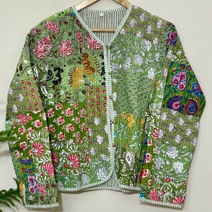 以下が含まれることがあります： ピンク、白、青の花柄が特徴の緑色のパッチワークジャケット。 ジャケットはVネックで、ストライプの生地で裏打ちされています。 木製のハンガーにかかっています。