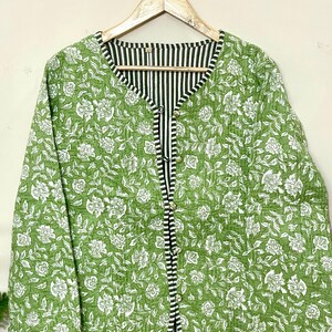 Puede incluir: Chaqueta verde con estampado floral y forro y ribete a rayas blancas y negras. La chaqueta es abotonada y tiene mangas largas. Cuelga de una percha de madera sobre un fondo neutro.