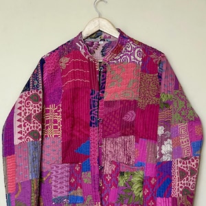 Puede incluir: Chaqueta patchwork en tonos rosas, morados y magenta, con cuello mandarín y cierre de botones. La chaqueta presenta una variedad de patrones y texturas de tela, incluyendo diseños florales y geométricos. Cuelga de una percha de madera.