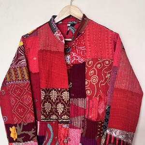 Op de afbeelding: Een patchwork jas in tinten rood, bordeaux en roze, met een opstaande kraag en knoopsluiting. De jas heeft een verscheidenheid aan stoffen vierkanten met patronen, waardoor een uniek en gestructureerd ontwerp ontstaat. Het hangt aan een houten hanger tegen een neutrale achtergrond.