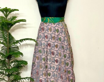 Puede incluir: Una falda cruzada con base rosa claro y un patrón repetitivo de líneas onduladas gris oscuro y motivos florales coloridos en rojo, verde y amarillo. La falda tiene una cintura ancha de color verde sólido y llega a media pierna en un maniquí.