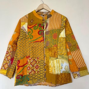 May include: A patchwork jacket in shades of yellow, orange, and green, with a mandarin collar and button closure. The jacket is made of various fabric patterns, including floral, geometric, and striped designs. The jacket is hanging on a wooden hanger.