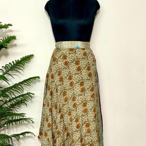 May include: A beige wrap skirt with a repeating floral pattern in shades of brown and green. The skirt is displayed on a black mannequin with a gold-colored belt. The skirt has a flowing, asymmetrical hemline.