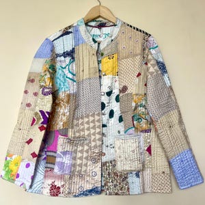 May include: A patchwork jacket with a mandarin collar and two front pockets. The jacket is made of various fabric scraps in beige, blue, yellow, and pink, sewn together in a quilted pattern. It hangs on a wooden hanger.