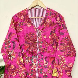 Puede incluir: Chaqueta acolchada fucsia con un estampado floral detallado en tonos amarillos, verdes y naranjas. La chaqueta tiene cuello en V y está forrada con una tela a rayas. La chaqueta de manga larga está colgada en una percha de madera.