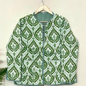 Puede incluir: Chaqueta reversible acolchada con un estampado ikat verde y blanco. La chaqueta tiene cuello redondo, dos bolsillos y forro a rayas. Cuelga de una percha de madera.