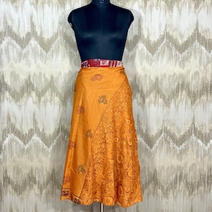 Könnte beinhalten: Ein orangefarbener Wickelrock mit aufwendigen Blumen- und Paisley-Mustern. Der Rock wird auf einer schwarzen Schaufensterpuppe mit einem roten und gemusterten Bund präsentiert. Der Rock besteht aus leichtem Stoff und hat eine fließende Silhouette.