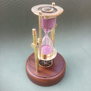 Könnte beinhalten: Eine Sanduhr mit Messingrahmen und Holzsockel. Die Glaskammer enthält rosa Sand. Auf der Oberseite der Sanduhr steht "SS LONDON 1917". Die Sanduhr ist ein dekorativer Artikel.