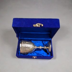 Pode incluir: Um cálice prateado com padrões florais ornamentados, apresentado numa caixa de veludo azul. O cálice tem um design detalhado na taça e haste, e a caixa tem um fecho dourado.