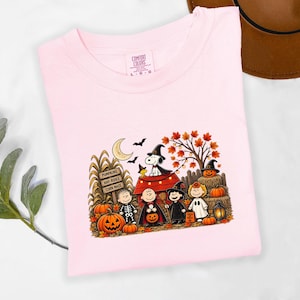 May include: A light pink t-shirt featuring a graphic print of Peanuts characters dressed in Halloween costumes. The design includes Snoopy on a red doghouse, pumpkins, a sign reading PUMPKINS HAY-RIDES CORN MAZE, and autumn leaves.