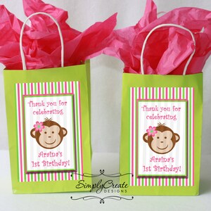 May include: Two green gift bags with pink and green striped labels. The labels feature a cartoon monkey with a pink flower and the text "Thank you for celebrating Araina's 1st Birthday!"