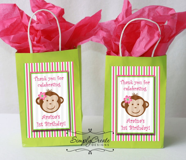 May include: Two green gift bags with pink and green striped labels. The labels feature a cartoon monkey with a pink flower and the text "Thank you for celebrating Araina's 1st Birthday!"