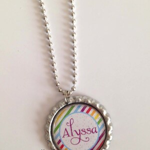 May include: A silver chain necklace with a round pendant. The pendant is a bottle cap with a rainbow striped design and the name "Alyssa" written in purple cursive.