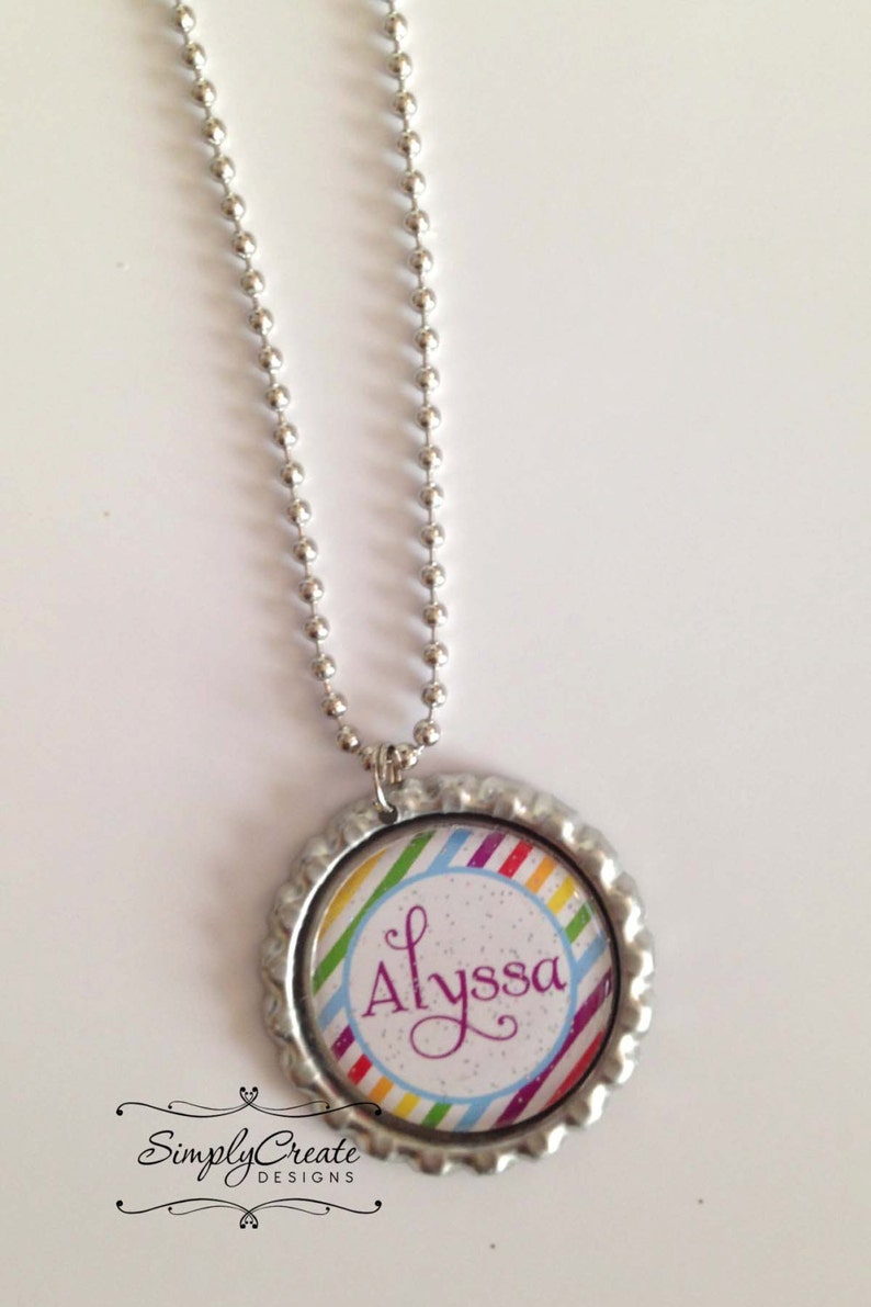 May include: A silver chain necklace with a round pendant. The pendant is a bottle cap with a rainbow striped design and the name "Alyssa" written in purple cursive.