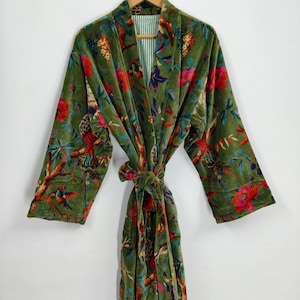 Może przedstawiać: Zielony aksamitny szlafrok z kwiatowym i ptasim wzorem. Szlafrok ma długie rękawy i wiązany pasek. Wewnętrzna podszewka jest w zielono-białe paski. Szlafrok wisi na drewnianym wieszaku na białym tle.