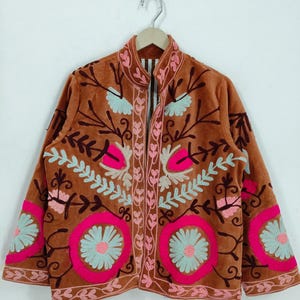 May include: A brown jacket with intricate floral embroidery in pink and light blue. The jacket features a high collar and long sleeves, with the floral design covering the front and sleeves. The jacket is hanging on a wooden hanger.