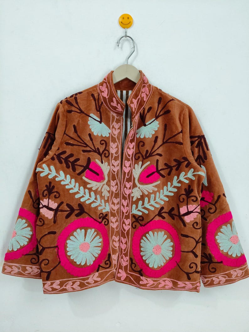 May include: A brown jacket with intricate floral embroidery in pink and light blue. The jacket features a high collar and long sleeves, with the floral design covering the front and sleeves. The jacket is hanging on a wooden hanger.
