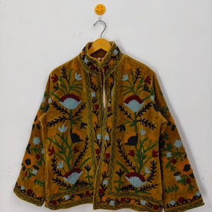 May include: Mustard velvet jacket with detailed floral embroidery in blue, red, green, and orange. The jacket has a high collar and long sleeves, hanging on a wooden hanger. The embroidery creates a textured pattern. The jacket is a unique piece of clothing.
