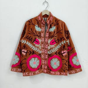 May include: A brown jacket with intricate floral embroidery in pink, light blue, and white. The jacket features a high collar and a full-length zipper. The embroidery is concentrated on the front, sleeves, and collar.