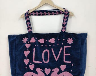 May include: A navy blue tote bag with pink embroidered details. The word "LOVE" is centered, with hearts, flowers, and two birds. The straps have pink embroidered leaf patterns. The bag is hanging on a wooden hanger.