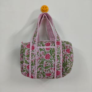 May include: A quilted tote bag with a floral pattern in pink, green, and white. The bag has pink and white striped handles and trim. The bag is hanging on a white wall.