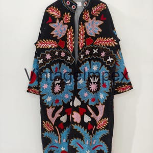 May include: A black long-line coat featuring vibrant multi-colored floral and leaf embroidery in shades of blue, red, pink, and white. The garment has a high collar, long sleeves, and a vertical striped lining visible at the neck opening.