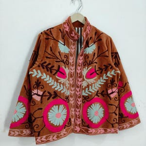 May include: A brown velvet jacket with intricate floral embroidery in pink, light blue, and brown. The jacket features a stand-up collar and is hanging on a wooden hanger against a white background.