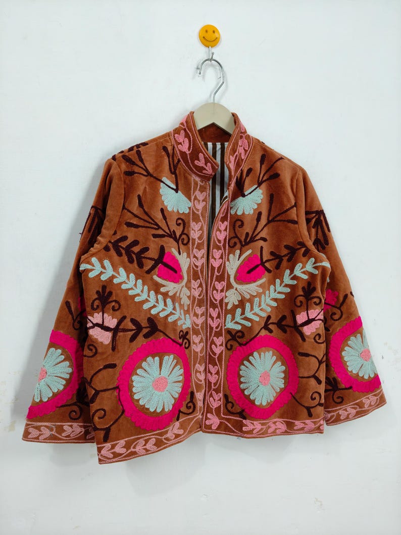 May include: A brown velvet jacket with intricate floral embroidery in pink, light blue, and brown. The jacket features a stand-up collar and is hanging on a wooden hanger against a white background.