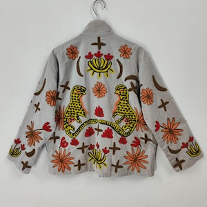 Puede incluir: Chaqueta gris claro con bordados coloridos. La parte trasera presenta dos figuras amarillas y negras con forma de leopardo enfrentadas, rodeadas de diseños florales naranjas y rojos y cruces marrones.