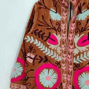 May include: A brown velvet jacket with intricate floral embroidery in pink, light blue, and cream. The jacket features a mandarin collar and long sleeves, showcasing a detailed, colorful design. The embroidery includes floral and leaf patterns.