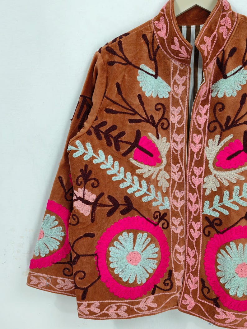 May include: A brown velvet jacket with intricate floral embroidery in pink, light blue, and cream. The jacket features a mandarin collar and long sleeves, showcasing a detailed, colorful design. The embroidery includes floral and leaf patterns.