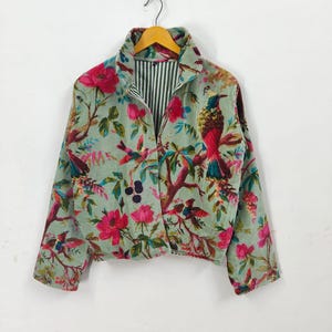 Peut inclure: Veste vert clair ornée d'un imprimé floral et d'oiseaux. La veste a un col, des manches longues et une fermeture boutonnée. La doublure intérieure est à rayures noires et blanches. La veste est suspendue à un cintre en bois.