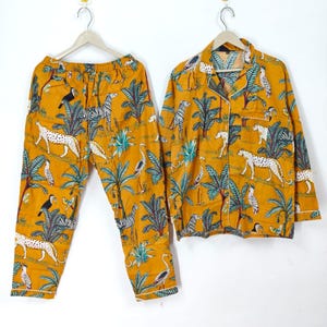 May include: Mustard yellow pajama set with a jungle animal print. The set includes a button-down shirt and pants, both featuring zebras, leopards, birds, and palm trees. White trim accents the edges of the shirt and pants.