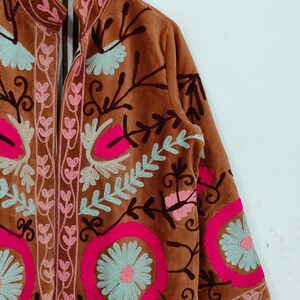 May include: A brown velvet jacket with intricate floral embroidery in pink, light blue, and dark brown. The jacket features a high collar and long sleeves, with detailed patterns around the edges and on the sleeves.