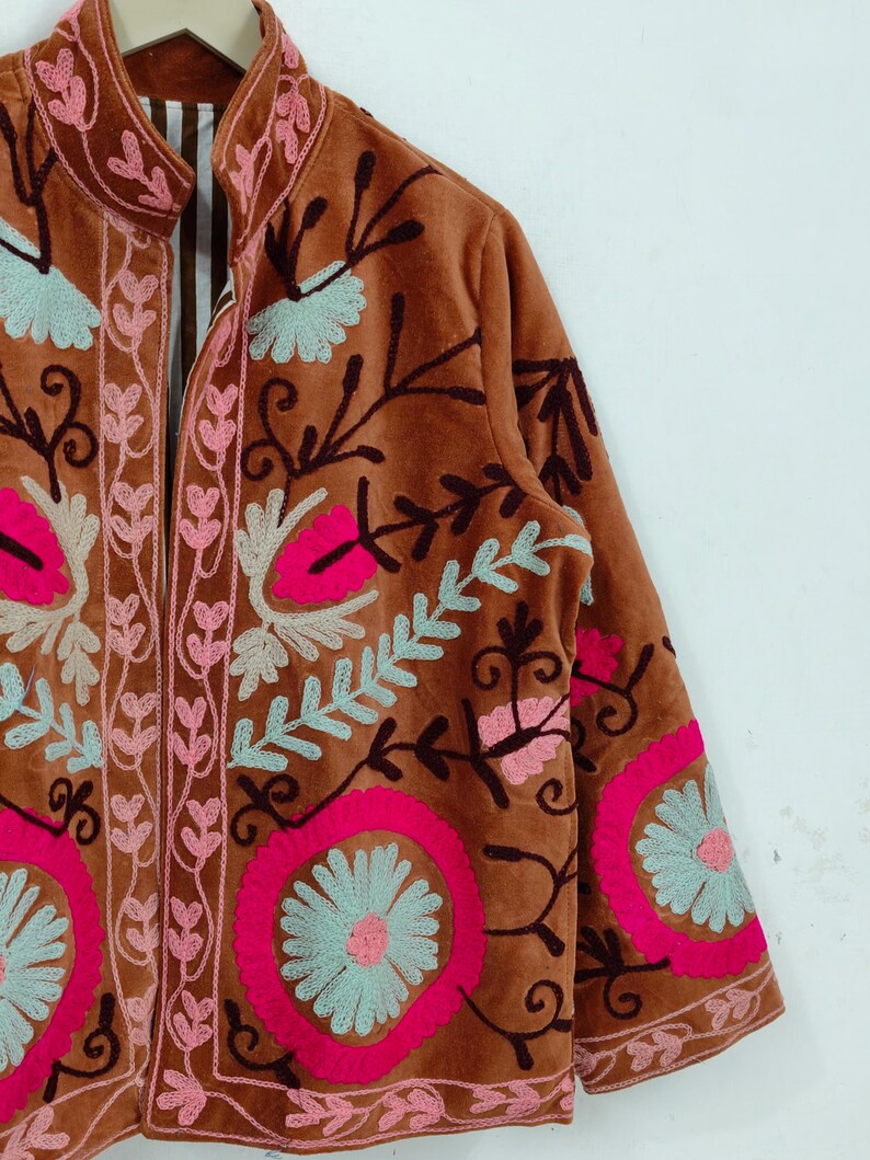 May include: A brown velvet jacket with intricate floral embroidery in pink, light blue, and dark brown. The jacket features a high collar and long sleeves, with detailed patterns around the edges and on the sleeves.