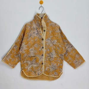 May include: A mustard yellow quilted jacket with a floral pattern in shades of gray and beige. The jacket has a high collar, a button-up front, and two front pockets. The jacket is hanging on a hanger.