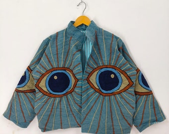 Könnte beinhalten: Türkisfarbene Jacke mit gestickten Augendesigns. Die Jacke zeigt große, stilisierte Augen in Blau-, Braun- und Goldtönen, die mit braunen Linien nach außen strahlen. Die Jacke hängt an einem Holzbügel vor weißem Hintergrund.
