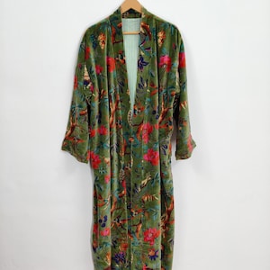 May include: An olive green velvet robe with a floral and bird design in red, pink, and blue. The robe is hanging on a wooden hanger. The inside lining is a green and white striped fabric. The background is a white wall.