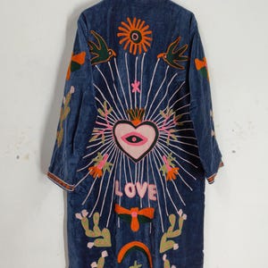 May include: A long, dark blue embroidered coat with a high collar. The coat features colorful embroidered designs, including a heart, sun, birds, and the word "LOVE". The embroidery is in shades of pink, orange, green, and white.
