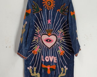 Puede incluir: Abrigo largo bordado azul oscuro con cuello alto. El abrigo presenta diseños bordados coloridos, incluyendo un corazón, sol, pájaros y la palabra "LOVE". El bordado es en tonos rosa, naranja, verde y blanco.
