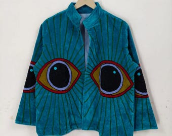 Könnte beinhalten: Türkisfarbene Jacke mit hohem Kragen und Reißverschluss. Die Jacke hat große Augendesigns auf der Vorderseite und den Ärmeln, mit schwarzen Pupillen, goldenen Iris und roten und lila Umrissen. Vertikale Linien strahlen von den Augen aus.