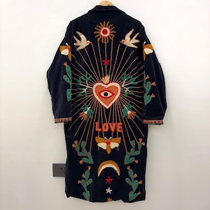 May include: A long, dark blue embroidered coat with a bohemian design. The back features a heart with an eye, the word "LOVE", birds, cacti, a sun, and stars. The coat has a relaxed fit and is likely made of a soft fabric.