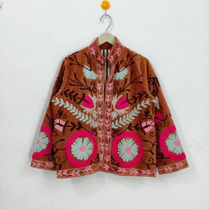 May include: A brown jacket with intricate floral embroidery in pink, light blue, and white. The jacket features a high collar and a zip-up front. The sleeves and front panels are adorned with large, circular floral designs.