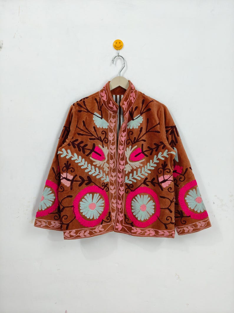 May include: A brown jacket with intricate floral embroidery in pink, light blue, and white. The jacket features a high collar and a zip-up front. The sleeves and front panels are adorned with large, circular floral designs.