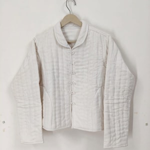 May include: Off-white quilted jacket with a rounded collar and long sleeves. The jacket features a button-up front and a diamond-patterned stitching design. The jacket is hanging on a wooden hanger.