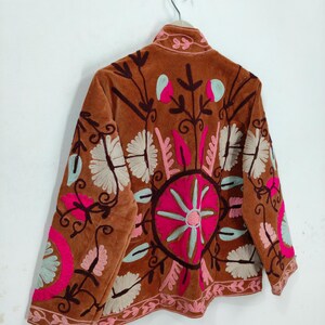 May include: A brown velvet jacket with intricate floral embroidery in pink, white, and teal. The jacket features a high collar and a detailed circular design on the back, showcasing a bohemian style.